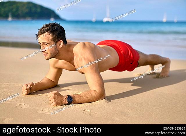 Athletic tanned man does a plank on the sand beach on the sunny background of the sea with white boats and the blue sky. He wears a red swim trunks and a dark...
