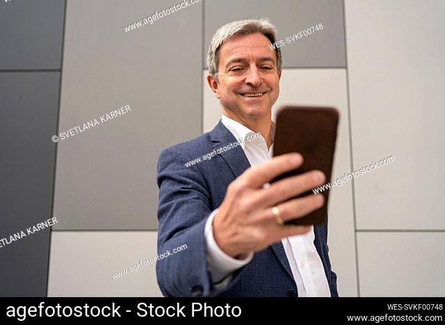 Happy mature businessman using smart phone in front of gray wall
