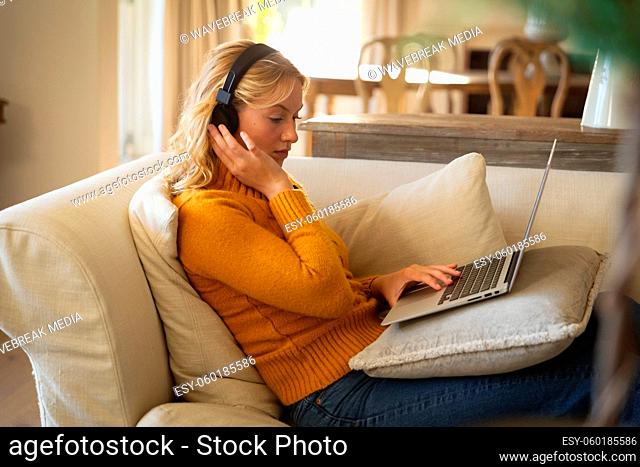 Caucasian woman relaxing on couch in living room wearing headphones and using laptop