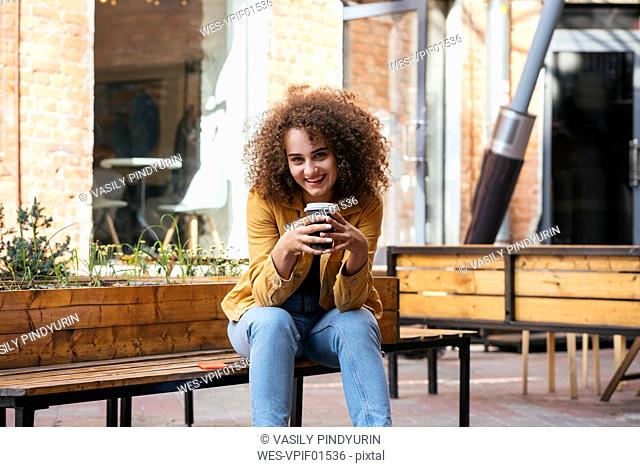 Portrait of smiling teenage girl sitting on bench with coffee to go