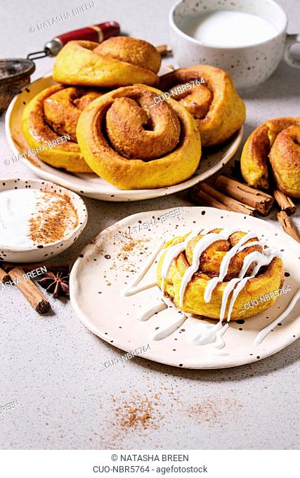 Homemade pumpkin cinnamon bun rolls sweet autumn baked dessert with cream cheese sauce in spotted ceramic plates with cinnamon sticks over white marble table