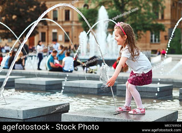 Little happy girl playing with the water in fountain in the center of town. Child catching a water stream going from a fountain