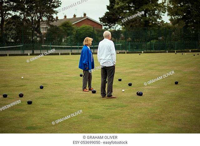 Two senior adults standing together in the middle of a bowling green, surrounded by bocce balls