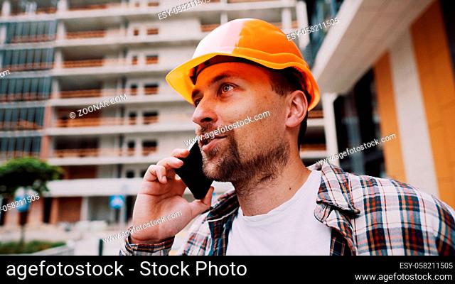 Portrait of young handyman making call while standing at construction area. Engineer talking on the phone on a construction site