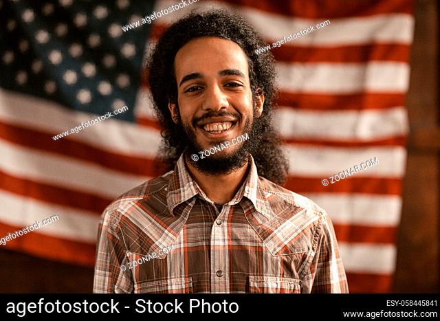 Young American Citizen Of Arab Nationality Laughs Cheerful, Against The Backdrop Of The American Flag In Office