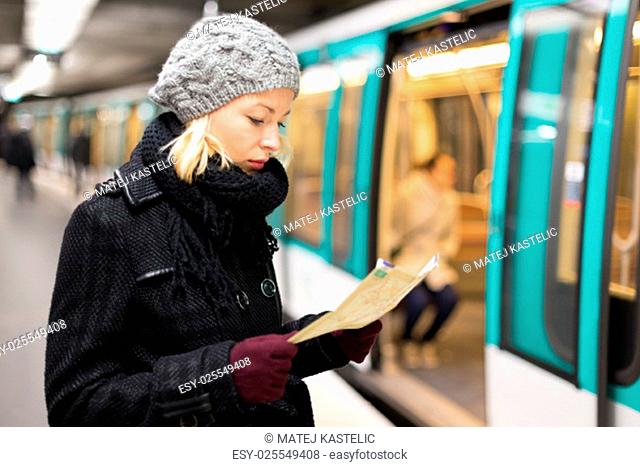 Casually dressed woman wearing winter coat, waiting on a platform for a train to arrive, orientating herself with public transport map