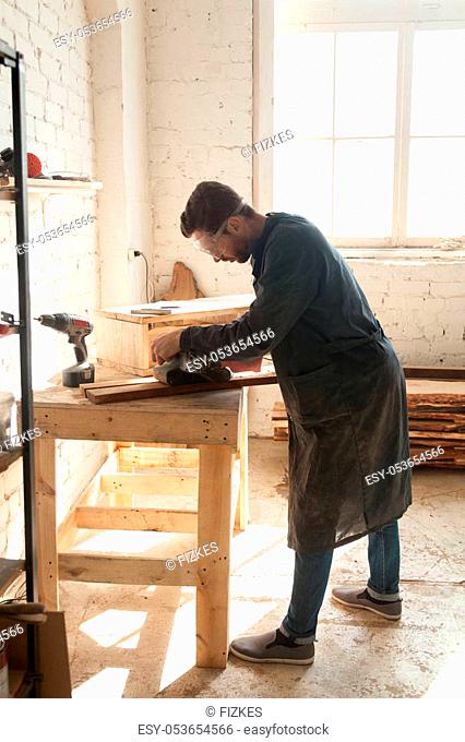 Carpenter polishes wooden boards with belt sanding machine while working in workshop. Young man choosing working profession at time of shortage skilled labor...