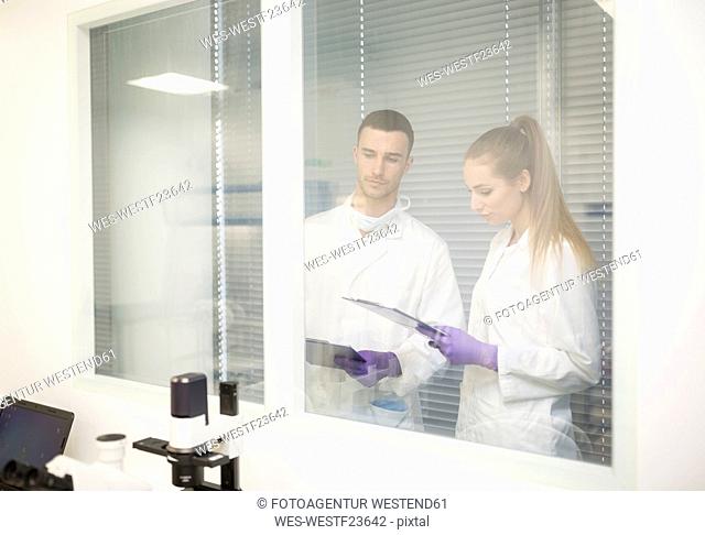 Man and woman in lab coats with clipboards behind glass pane