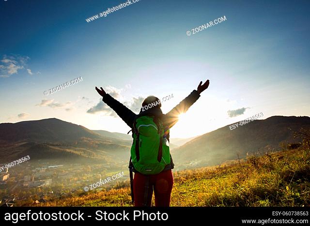 Portrait of young woman with backpack, standing and looking at the rising sun while hiking in mountains. Woman escaped from the routine to reconnect with nature
