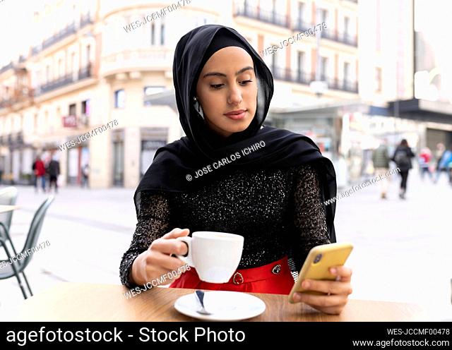 Portrait of young beautiful woman wearing black hijab using smart phone at sidewalk cafe