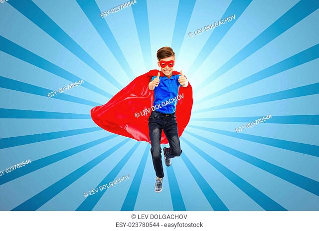 happiness, freedom, childhood, movement and people concept - boy in red super hero cape and mask flying in air and showing thumbs up over blue burst rays...