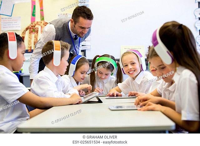 Group of children wearing colourful wireless headsets while working on digital tablets, the teacher can be seen supervising the students in the classroom