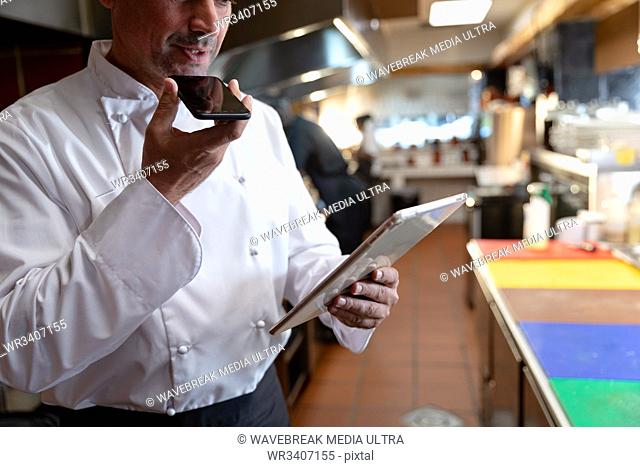 Front view close up of a middle aged Caucasian male chef holding and talking on a smartphone while he looks at a tablet computer he holds in the other hand in a...