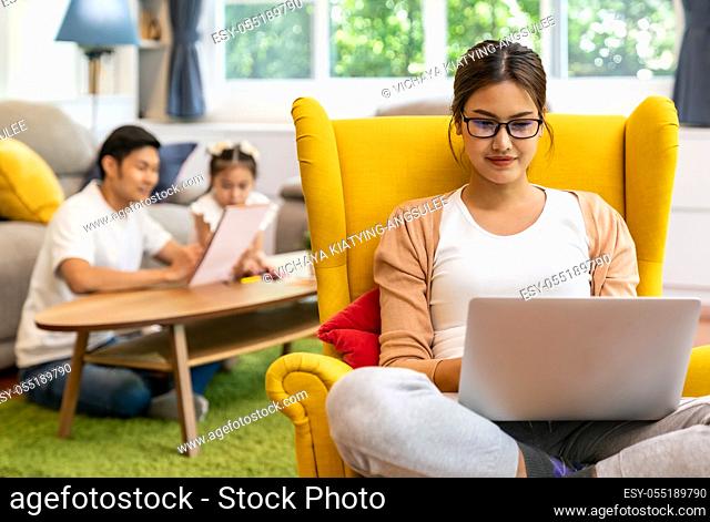 Asian working mom work from home in living room with background of father and daughter reading tale story book while city lockdown