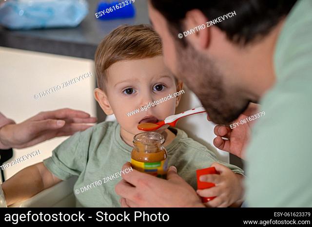 Breakfast for a babyboy. Young father feeding his cute baby son and looking funny