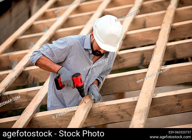 The male worker in a protective helmet and gloves works on the roof using a screwdriver. He finishes the wooden frame on the roof