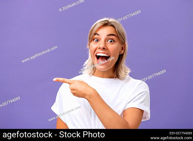 Waist-up shot of joyful thrilled attractive and emotive young blonde female with tanned skin in white casual t-shirt smiling and laughing happily pointing left...