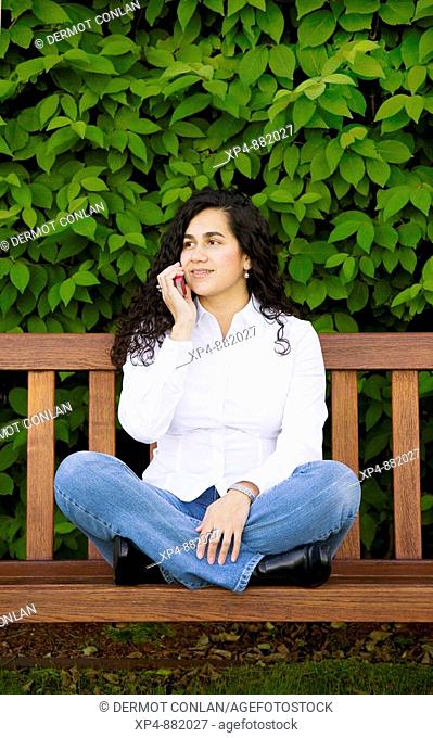 Young Colombian woman with long black hair white shirt and blue jeans, sits cross-legged on a wooden bench while talking on a mobile phone