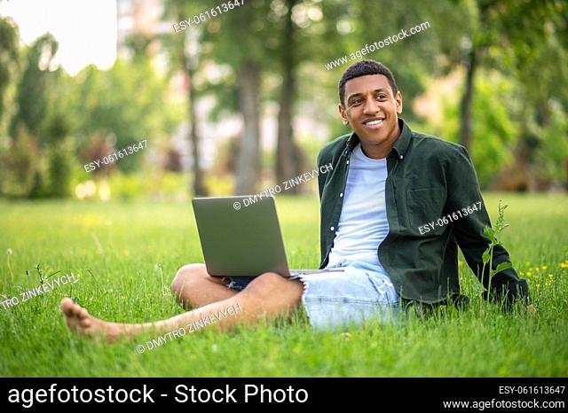 Solitude. Dark-skinned smiling guy barefoot with laptop sitting on grass in park looking thoughtfully to side