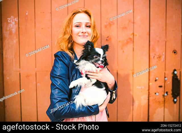 Young caucasian red-haired woman holding a dog of Chihuahua breed on her hands on a wooden background. The theme of love for animals, protection of pets
