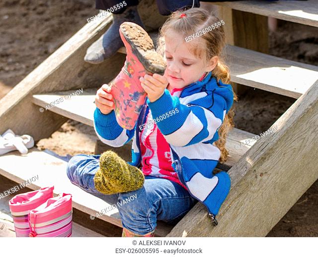 Four-year girl puts on boots sitting on the porch of a house in the village