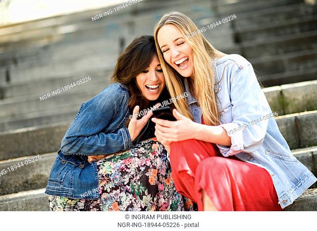 Two girls looking at some funny video on their smart phone outdoors, sitting on urban steps. Female friends girls laughing