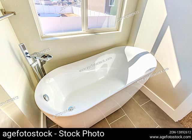 Bathroom interior with a gleaming bathtub against white wall and gray floor. The room is well lit by the sun that streams in through the large sliding window