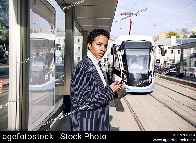 Young businesswoman with mobile phone waiting for bus at stop on sunny day