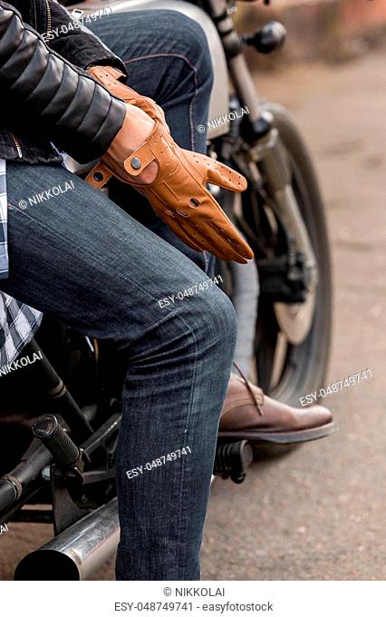 Close-up of a handsome rider biker man hand puts on a glove while sit on classic style cafe racer motorcycle. Bike custom made in vintage garage