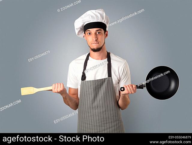 Young bearded cook portrait with kitchen tools and empty wallpaper