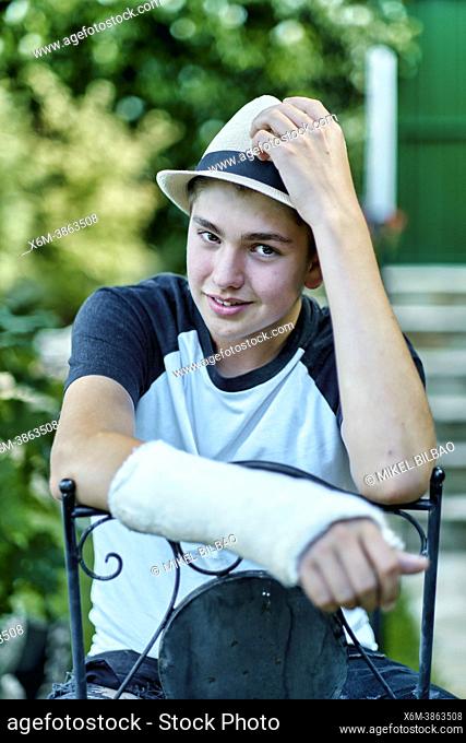 Portrait of young caucasian boy with a broken and cast arm wearing a hat and sitting in a chair outdoor in a garden. Lifestyle concept