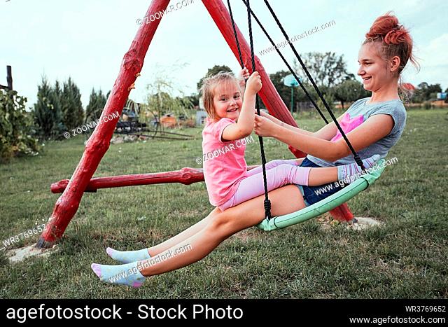Teenage girl playing with her younger sister in a home playground in a backyard. Happy smiling sisters having fun on a swing together on summer day