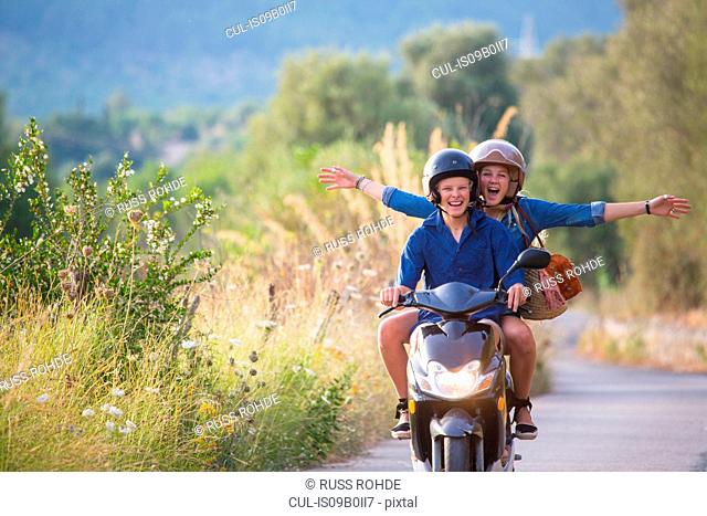 Young woman riding pillion on moped with arms open on rural road, Majorca, Spain