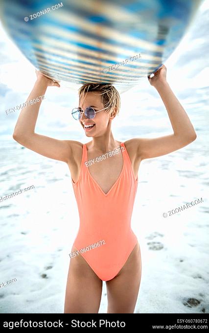 Smiling blonde girl with short hairstyle stands on the beach on the background of the sea and cloudy sky. She wears orange swimsuit with sunglasses and holds...