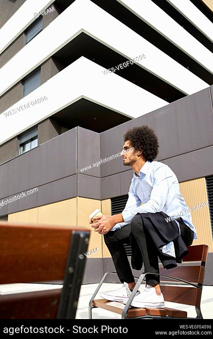 Afro man with coffee cup looking away while sitting on chair in city