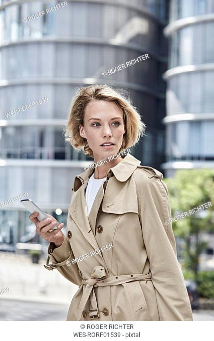 Germany, Duesseldorf, portrait of blond young businesswoman with smartphone wearing beige trenchcoat