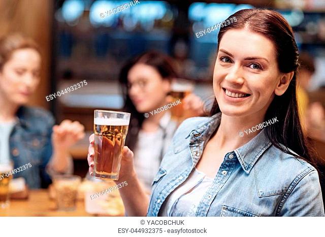 Just Friday. Positive delighted woman holding bocal of beer in right hand looking straight on camera while having weekend party