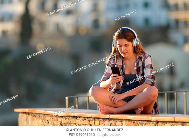 Happy teen listening to music sitting on a ledge in a town outskirts on holiday