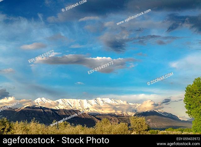 Scenic nature view with majestic snow peaked mountain under cloudy blue sky. Trees with lush green leaves can be seen in foreground on this sunny day