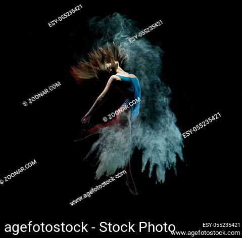 Slim young woman in blue leotard jumping and shaking hair while dancing in cloud of dust against black background