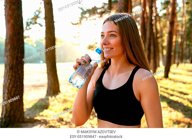 Outdoor photo of young sportive woman drinking a water from bottle after running in the park. Dressed in black tank top