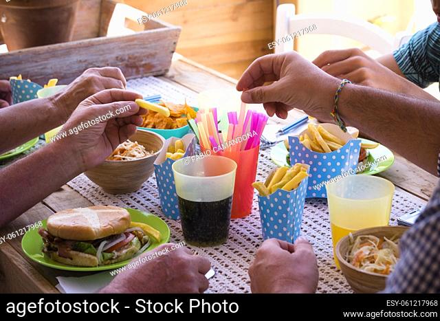 Close up of caucasian friends hands taking fast food hamburger and frech fries from the table - group of unrecognizable people eating and having fun together