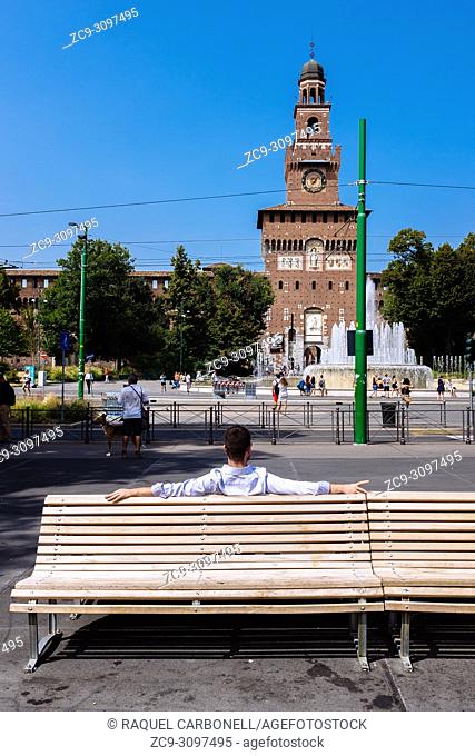 Young man sit on a public bench looking to Piazza Castello square fountain with the Torre del Filarete in Sforza Castle at back, Milan, Lombardy, Italy