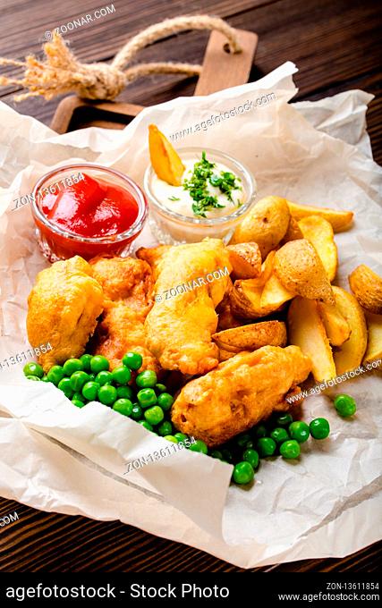 Close-up of British traditional fast food fish and chips with assorted dips, peas, on paper, rustic brown wooden background