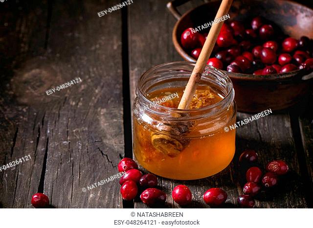 Open glass jar of liquid honey with honeycomb and honey dipper inside and fresh cranberries in vintage bowl over old wooden table. Dark rustic style