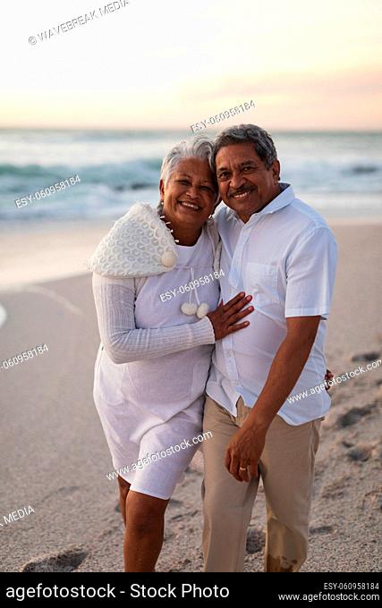 Portrait of smiling senior biracial couple embracing at beach against sky during sunset