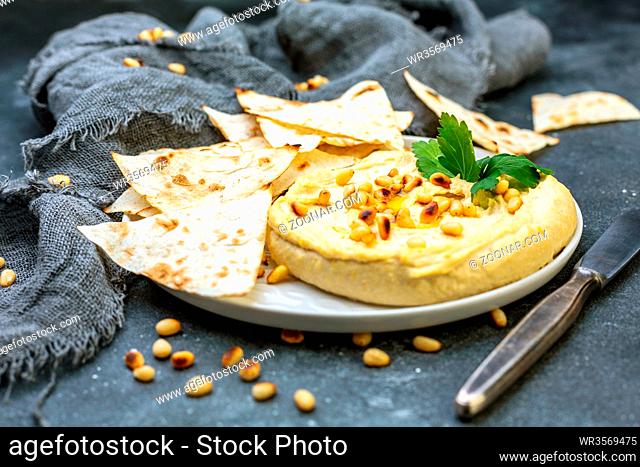 Plate of homemade traditional hummus, pieces of toasted homemade pita bread and pine nuts on a textured dark table, selective focus