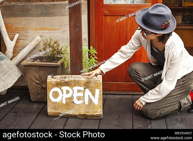 Japanese woman wearing hat and glasses kneeling in front of a leather shop, holding Open sign