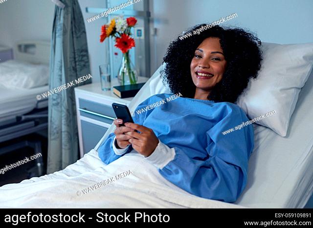 Portrait of smiling african american female patient lying in hospital bed using smartphone