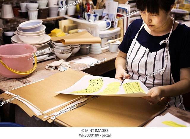 Woman sitting in a Japanese porcelain workshop, cutting out design patterns with a pair of scissors
